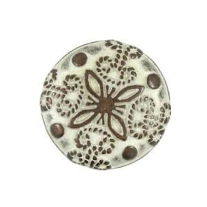 May include: A round, decorative button with a cream-colored background and a brown floral design in the center. The button has a distressed, vintage look, with small brown dots around the edge.