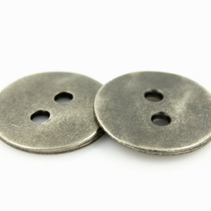 Puede incluir: Dos botones de metal plateados con dos agujeros cada uno. Los botones tienen una superficie ligeramente texturizada.