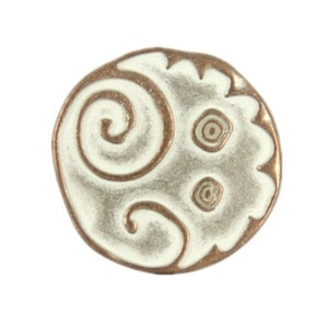 May include: A circular decorative bead with a white central design and a bronze-coloured border. The white area features a spiral pattern and two small concentric circles. The bead has a scalloped edge.