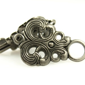May include: A silver-toned metal clasp with an ornate design featuring multiple loops and swirls. The clasp is designed to be used for jewelry making or other craft projects.