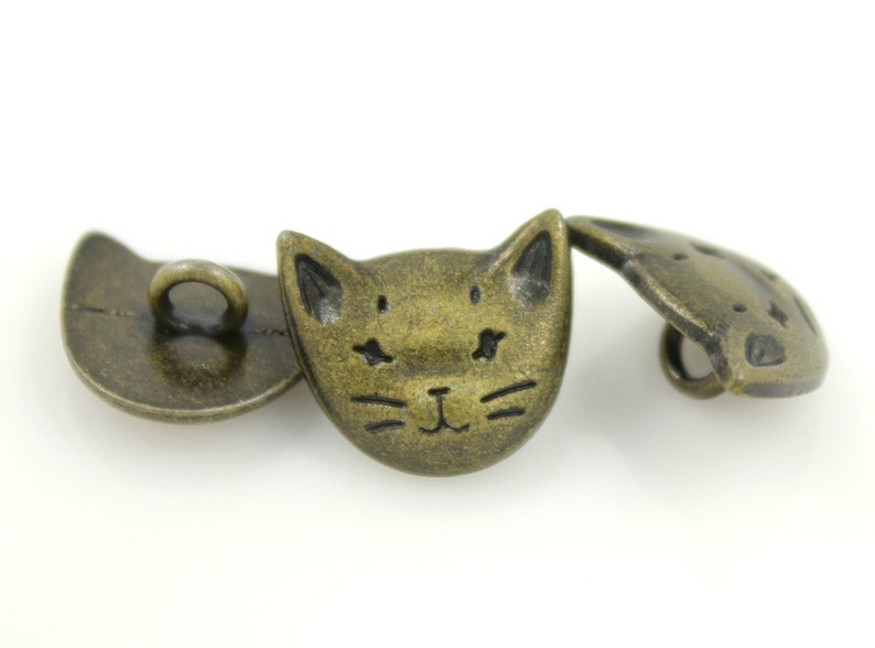 May include: Three antique gold-toned metal buttons with a cat face design. The buttons have a hole for sewing.