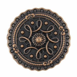 May include: A round, copper-colored button with a black, intricate design. The button has a raised, circular border with a beaded edge.