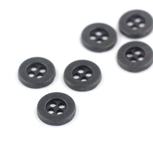 Pode incluir: Seis botões de plástico preto com quatro furos cada. Os botões estão dispostos de forma dispersa sobre um fundo branco.