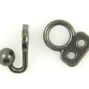 Puede incluir: Dos fornituras de joyería de metal plateado antiguo. Una es un gancho con un extremo de bola, y la otra es un anillo con una base rectangular y dos agujeros. Estas fornituras se utilizan probablemente para la fabricación de joyas o manualidades.