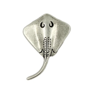 Puede incluir: Un amuleto de metal plateado con forma de raya. El amuleto tiene un diseño detallado con un cuerpo texturizado y una cola larga.