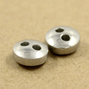 Puede incluir: Dos botones de metal plateado con dos agujeros cada uno. Los botones son redondos y tienen un acabado liso y brillante.