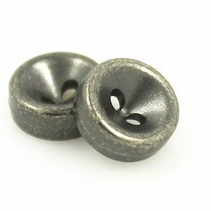 Può includere: Due bottoni in metallo color argento con un bordo rialzato e tre fori. I bottoni sono rotondi e hanno un centro leggermente concavo.