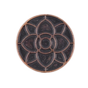Op de afbeelding: Een ronde, koperkleurige metalen knoop met een zwart, ingewikkeld ontwerp van een gestileerde bloem met een cirkelvormig midden.