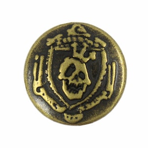Peut inclure: Un bouton rond en métal doré avec un motif de crâne et d'os croisés. Le crâne est entouré d'une couronne de feuilles et les os croisés sont formés par deux épées croisées.