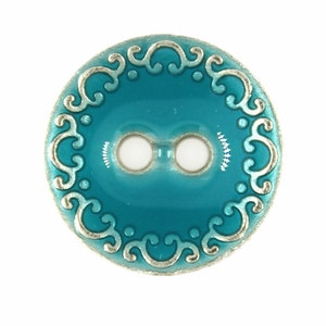 Puede incluir: Un botón azul turquesa con un borde metálico plateado y un diseño decorativo de desplazamiento. El botón tiene dos agujeros.