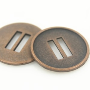 Puede incluir: Dos hebillas redondas de metal de color cobre con una abertura de doble ranura. Las hebillas tienen una superficie ligeramente texturizada.