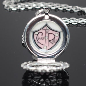 May include: Silver locket necklace with a pink and white shield design featuring the text "The CTR" and "those who rise" on the front. The locket is open, revealing a delicate, intricate design.