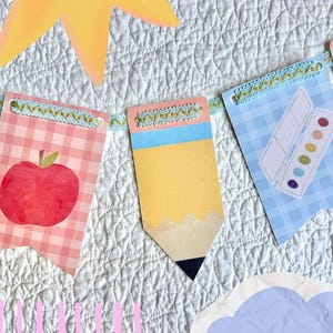 Peut inclure: Une guirlande décorative composée de fanions avec des illustrations sur le thème de l'école, dont une pomme rouge sur fond vichy rose, un crayon jaune et une boîte d'aquarelle sur fond vichy bleu, le tout relié par une bordure bleue.