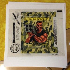 May include: A vintage trading card featuring a cartoon illustration of Arnold Schwarzenegger as John Matrix from the movie Commando. The card is green, black, and white with the text "COMMANDO" in large, bold letters. The card includes the text "10 Movie Photo Cards, 1 Sticker, 1 Stick Bubble Gum".