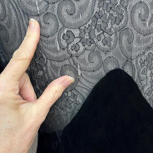 以下が含まれることがあります： 黒い衣服のクローズアップ。複雑なレースのディテールが特徴です。レースは花柄と渦巻き模様です。レースの下には、黒いベルベット生地の部分があります。画像は、素材の質感とパターンを強調しています。