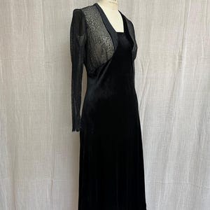 以下が含まれることがあります： 黒いベルベットのドレスと、透け感のあるレースのボレロジャケット。 ドレスはスクエアネックで、スカートは流れるようなデザインです。 ボレロは長袖で、繊細なレースのディテールが特徴です。 ドレスはマネキンに着せられています。