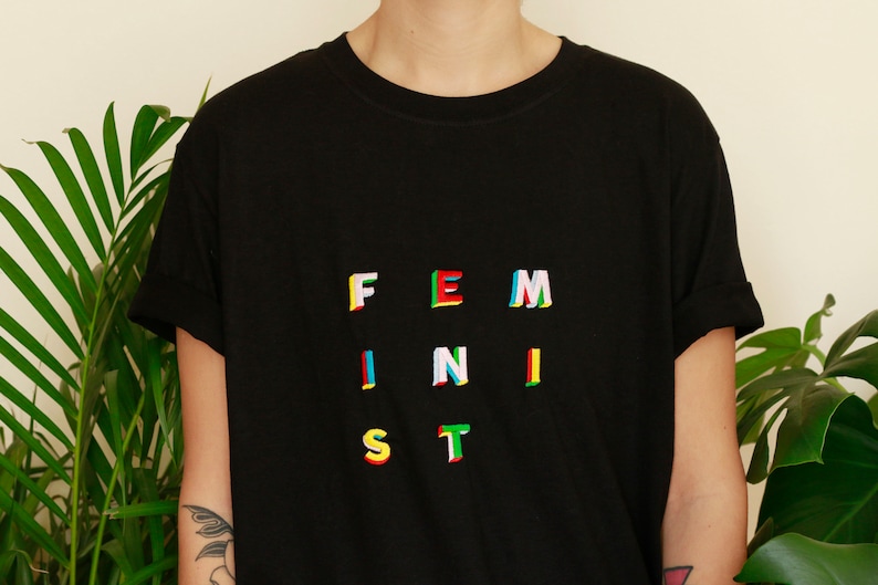 Puede incluir: Una camiseta negra con la palabra "FEMINIST" escrita en letras coloridas y multicapa.