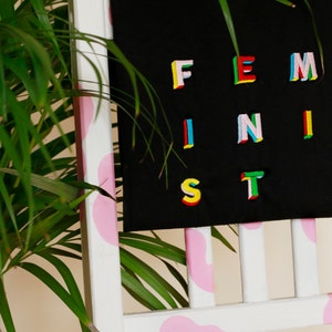 Puede incluir: Una camiseta negra con la palabra "FEMINIST" escrita en letras coloridas y multidimensionales. La camiseta est&aacute; colgada en una silla blanca con detalles rosas.