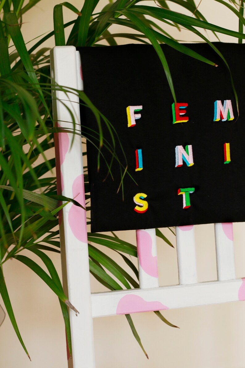 Puede incluir: Una camiseta negra con la palabra "FEMINIST" escrita en letras coloridas y multidimensionales. La camiseta est&aacute; colgada en una silla blanca con detalles rosas.