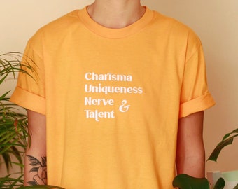 Puede incluir: Una camiseta amarilla con el texto "Charisma Uniqueness Nerve & Talent" impreso en blanco.