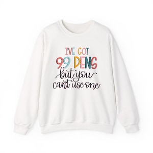 May include: A white long-sleeve crewneck sweatshirt featuring the colorful text I'VE GOT 99 PENS but you can't use one. The words 99 PENS are printed in large, multi-colored block letters, while the remaining text is in a black script font.
