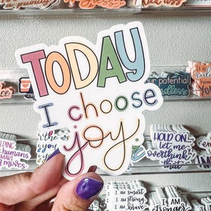 Op de afbeelding: Een witte sticker met de woorden "TODAY I choose joy" in kleurrijke bubbelletters en een cursief lettertype. De sticker wordt vastgehouden door een hand met een paars gelakte nagel. Andere stickers met motiverende zinnen zijn zichtbaar op de achtergrond.