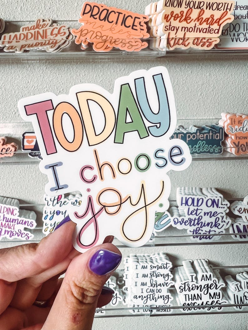Op de afbeelding: Een witte sticker met de woorden "TODAY I choose joy" in kleurrijke bubbelletters en een cursief lettertype. De sticker wordt vastgehouden door een hand met een paars gelakte nagel. Andere stickers met motiverende zinnen zijn zichtbaar op de achtergrond.
