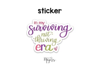 May include: Purple and green sticker with the text "in my surviving not thriving era" with a pink heart. The sticker is from "remarkable Meyers".