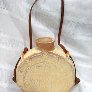 May include: A round, beige ceramic canteen with a glossy finish and a central embossed circular design. The vessel features a narrow neck and is fitted with a thin, brown leather strap threaded through two side loops for carrying purposes.