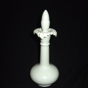 May include: A white ceramic perfume bottle with a bulbous base and a long, slender neck. The bottle is topped with a decorative stopper featuring a floral design. The background is solid black.