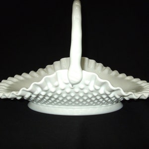 May include: A white hobnail basket with a handle. The basket has a ruffled edge and a textured surface. The handle is curved and attached to the basket. The basket is on a black background.