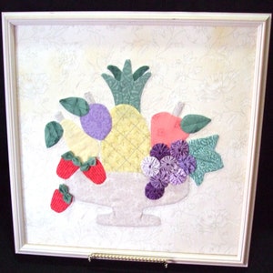May include: Framed fabric art featuring a still life of various fruits. The artwork includes a pineapple, strawberries, grapes, and other fruits in a white bowl. The frame is white, and the background is a floral pattern.