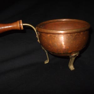 May include: A small copper pot with a wooden handle and three brass feet. The pot is on a black background.