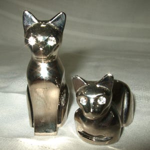 May include: A pair of polished silver-toned metal cat figurines. The taller cat stands upright, while the smaller cat is in a seated position. Both cat figures feature clear, sparkling rhinestone eyes that catch the light, creating a reflective surface.