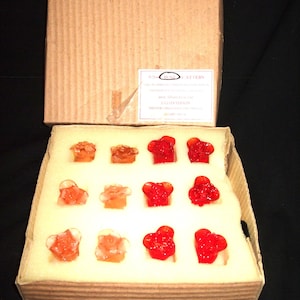 May include: A box of 12 glass rose bud ornaments in red and clear glass. The ornaments are arranged in a grid pattern in a foam-lined box. The box is labeled "Rose Attar" with the text "Handcrafted Glass Ornaments by Lily Vernon".