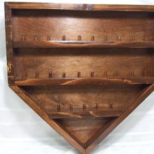 May include: A dark brown stained wooden display rack shaped like a home plate baseball base. The rack features three horizontal shelves, each equipped with small wooden pegs for organizing items, and a pointed bottom edge design.