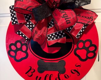 May include: A red wooden door hanger with a black "G" logo, paw prints, and the word "Bulldogs". The hanger is decorated with a large bow featuring red and black ribbons with the word "Georgia".