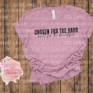 May include: A dusty rose t-shirt with the text "CHOSEN FOR THE HARD, built for the beautiful" in black. The shirt is made of soft material and is suitable for casual wear. The shirt is a crew neck.