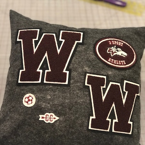 May include: A gray pillow with two large maroon felt letters "W" and a smaller maroon felt patch with the text "3 Sport Athlete" and a wolf graphic. There is also a small maroon felt arrow with the letters "CC" and a white soccer ball.