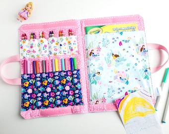 May include: Pink fabric art supply organizer with multiple pockets. Features pockets for crayons and markers, and a larger pocket with a mermaid print. Includes a handle for easy carrying. A small doll is visible in the upper left corner.