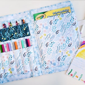 May include: A blue and white fabric carrying case with a floral pattern. The case has pockets for crayons and markers and a large pocket for a book. The book is a 3-in-1 activity puzzle book by Crayola.