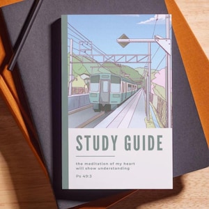 May include: A stack of notebooks and a pencil on a wooden surface. The top notebook is black, and the one beneath it is orange. A study guide with a train illustration is on top, with the text "STUDY GUIDE". A vase with purple flowers is in the background.