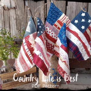 Puede incluir: Tres decoraciones de pared de tela hechas a mano con temática de la bandera estadounidense con diseños de patchwork rojo, blanco y azul. Las banderas se muestran en palos de madera y se apoyan contra un fondo de madera rústico. El texto "Country Life is Best PDF Pattern" es visible en la parte inferior de la imagen.