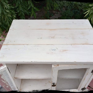 May include: A small, white, distressed wooden cabinet with a single door and a wire mesh front. The cabinet has a shelf inside and a flat top. The door is slightly ajar, revealing the interior. The cabinet is painted white with visible wood grain.