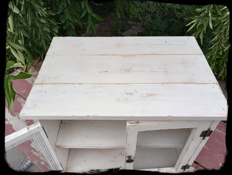 May include: A small, white, distressed wooden cabinet with a single door and a wire mesh front. The cabinet has a shelf inside and a flat top. The door is slightly ajar, revealing the interior. The cabinet is painted white with visible wood grain.