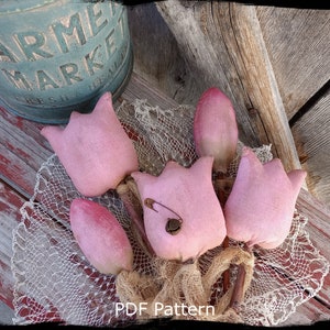 Peut inclure: Un patron PDF pour réaliser un ensemble de cinq tulipes en tissu rose. Les tulipes sont disposées sur une surface en bois rustique avec un morceau de dentelle et de toile de jute. Le texte "Country Life is Best" et "PDF Pattern" est visible sur l'image.