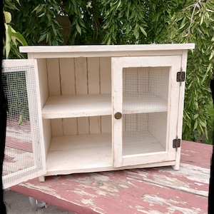 May include: A small, off-white cabinet with a distressed finish. The cabinet has a single door with wire mesh and a shelf inside. The door is open, revealing the interior. The cabinet is sitting on a red, weathered wooden surface.