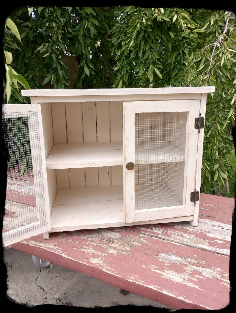 May include: A small, off-white cabinet with a distressed finish. The cabinet has a single door with wire mesh and a shelf inside. The door is open, revealing the interior. The cabinet is sitting on a red, weathered wooden surface.