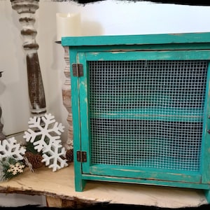 May include: Teal green wooden cabinet with a wire mesh door and a shelf inside. The cabinet has a distressed finish and a rustic look.