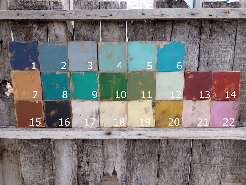 May include: A collection of twenty-two small, square wooden blocks, each painted a different color. The colors range from dark blue to light pink, with numbers 1 through 22 printed on each block. The blocks are arranged on a weathered wooden surface.